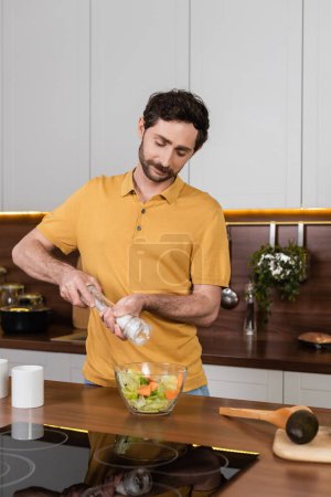 Hombre barbudo sosteniendo molino de sal mientras sazona ensalada en la cocina 