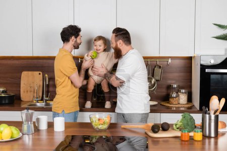 Cheerful gay couple holding toddler daughter with apple in kitchen 