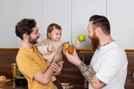 Pareja gay sosteniendo a su hija y comida para bebés en la cocina 