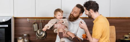 Photo for Smiling gay couple holding baby food near toddler daughter in kitchen, banner - Royalty Free Image