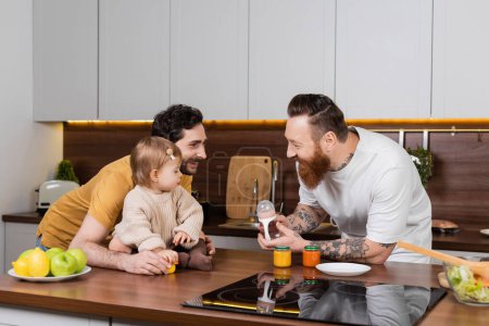 Gay couple holding baby bottle near toddler daughter in kitchen 