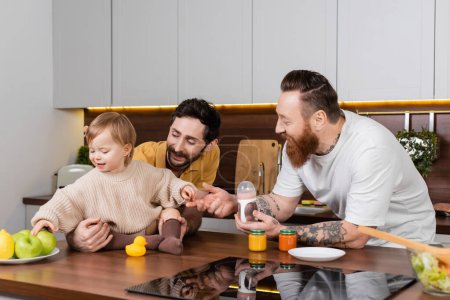 Smiling same sex couple holding baby bottle near toddler daughter in kitchen 