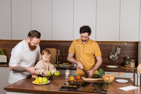Foto de Gay man cooking near partner and toddler daughter in kitchen - Imagen libre de derechos