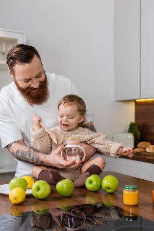Téléchargez les photos : Bearded man holding baby bottle near daughter with spoon and fruits in kitchen - en image libre de droit