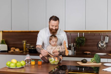 Tattooed father smiling and mixing salad near toddler daughter in kitchen 