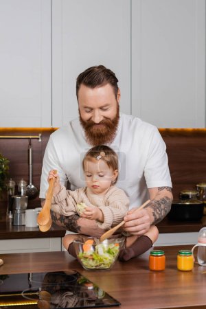 Smiling father cooking salad near daughter and baby food in kitchen 