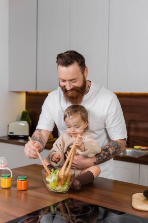 Foto de Bearded dad cooking fresh salad near toddler daughter in kitchen - Imagen libre de derechos