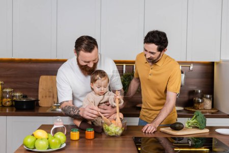 Foto de Gay man cooking salad near toddler daughter and partner in kitchen - Imagen libre de derechos