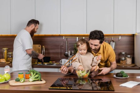 Gay man holding baby daughter with spoon near fresh salad in kitchen 