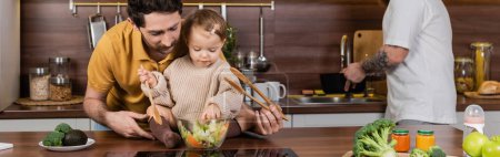 Homosexual parent holding daughter with spoon and fresh salad in kitchen, banner 