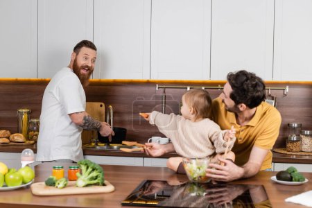 Foto de Excited gay man looking at daughter with vegetable near partner and fresh salad in kitchen - Imagen libre de derechos