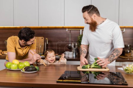 Lächelndes Kleinkind mit Löffeln in der Nähe von Vätern beim Kochen in der Küche 