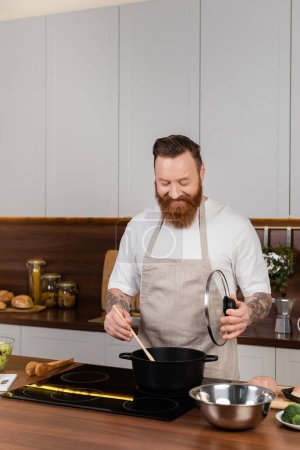 Hombre barbudo sonriendo y cocinando en olla en la cocina moderna 