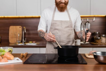 Vista recortada del hombre barbudo cocinando en olla cerca de la ensalada en la encimera en la cocina 