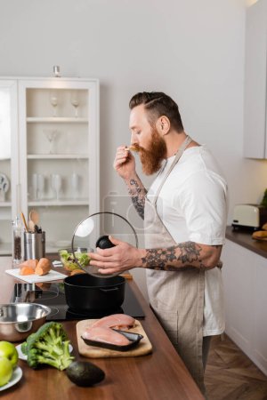 Side view of tattooed man in apron tasting food while cooking in kitchen 