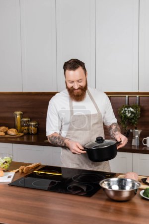 Homme barbu souriant tenant le pot près de la cuisinière et de la nourriture dans la cuisine 