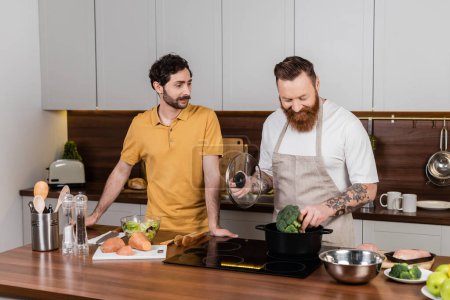 Hombre gay tatuado poniendo brócoli en la olla mientras cocina cerca de su pareja en la cocina 