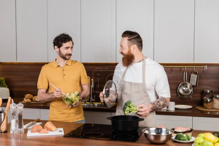 Homosexual man holding salad near partner cooking broccoli in kitchen 