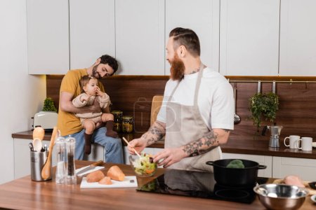 Gay father holding baby daughter while partner cooking in kitchen 