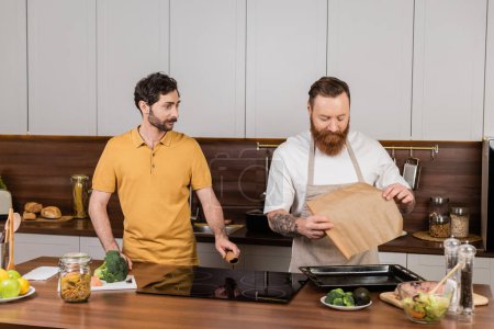 Téléchargez les photos : Gay man holding baking paper near raw chicken fillet and partner in kitchen - en image libre de droit