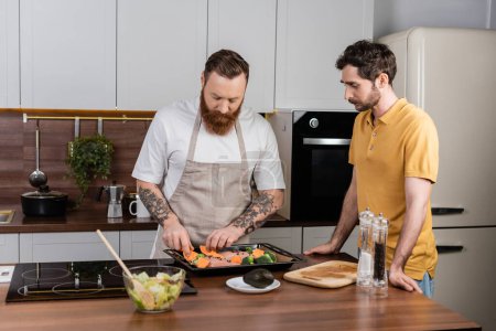 Gay man putting vegetables and chicken fillet on baking sheet near partner in kitchen 