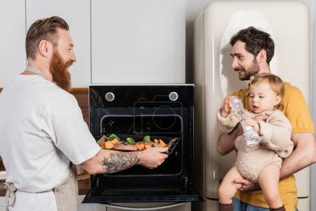 Smiling gay man putting food in oven near partner holding baby daughter in kitchen 