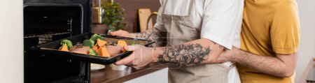 Geschnitten Ansicht der Homosexuell Mann umarmt Ehemann setzen Essen in den Ofen, während Kochen in der Küche, Banner 