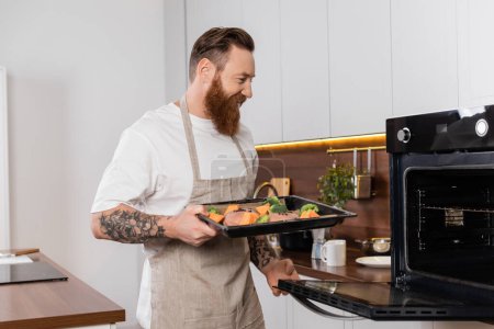 Foto de Sonriente hombre tatuado sosteniendo bandeja para hornear con carne y verduras cerca del horno en la cocina - Imagen libre de derechos