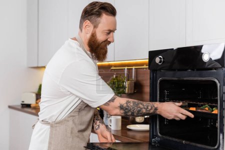 Lächelnder tätowierter Mann legt beim Kochen zu Hause Essen in den Ofen 