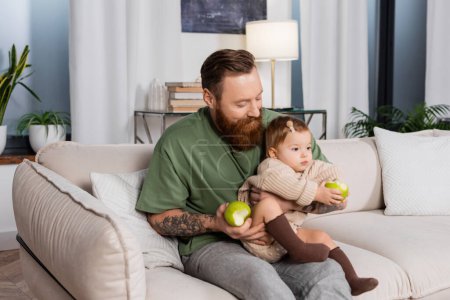 Bearded father holding apple and toddler daughter on couch at home 