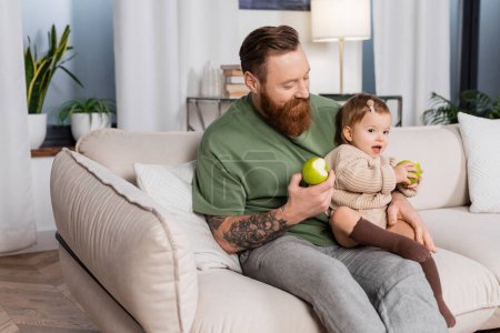 Tattooed man holding apple and baby daughter on couch at home 