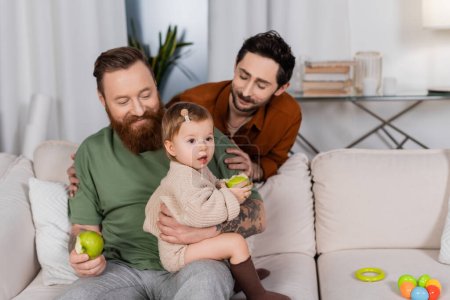 Smiling gay couple looking at baby daughter with apple on couch at home 