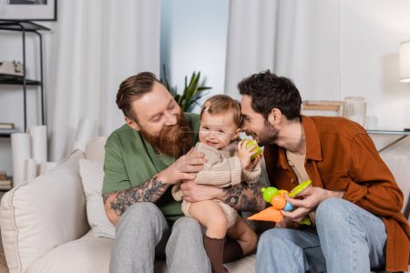 Happy gay couple holding baby girl with apple in living room 