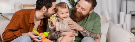 Photo for Positive same sex parents looking at baby daughter with apple at home, banner - Royalty Free Image