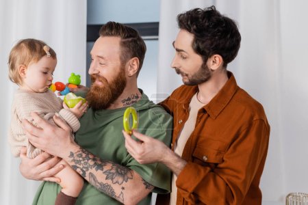 Smiling gay couple holding toys and baby girl with apple at home 