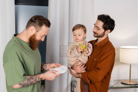 Gay man holding diaper near partner with baby daughter and apple at home 