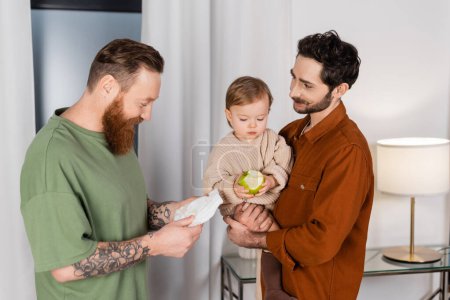 Positive gay couple holding baby daughter and diaper at home 