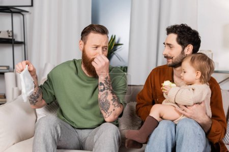Disgusted gay man plugging nose while holding diaper near partner with baby daughter at home 