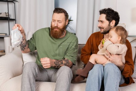 Disgusted gay parents holding diaper and baby girl at home 