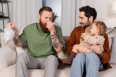 Padres homosexuales disgustados sosteniendo pañales cerca de marido e hija bebé en la sala de estar 