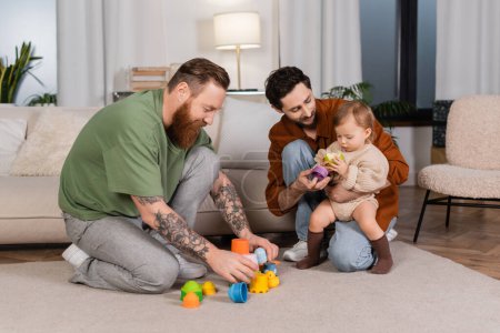 Same sex parents holding toys near baby girl with apple at home 