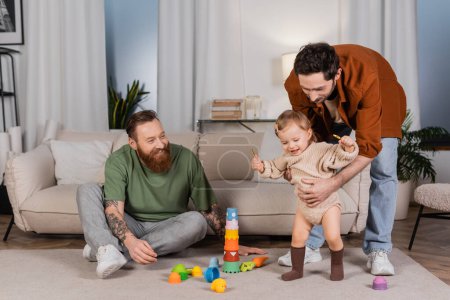 Positive gay parents playing with baby daughter near toys at home 