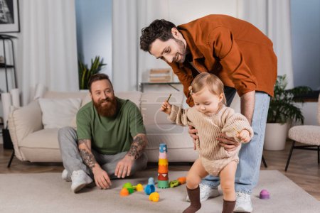 Smiling gay man holding baby daughter near partner and toys at home 
