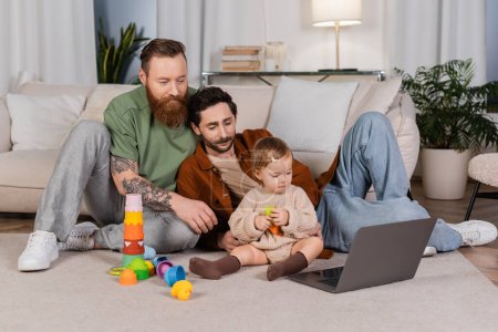 Gay couple looking at laptop near baby daughter with toys at home 