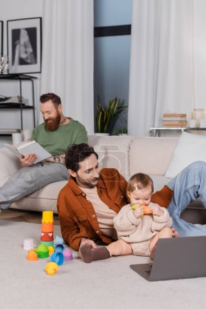 Gay parent sitting near baby daughter with toy and laptop in living room 