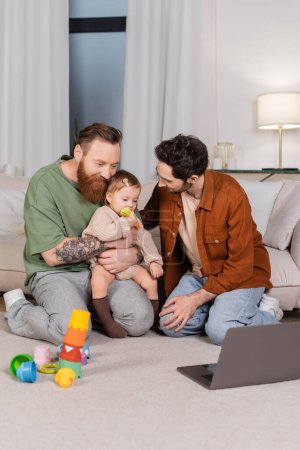 Gay couple hugging daughter near toys and laptop at home 