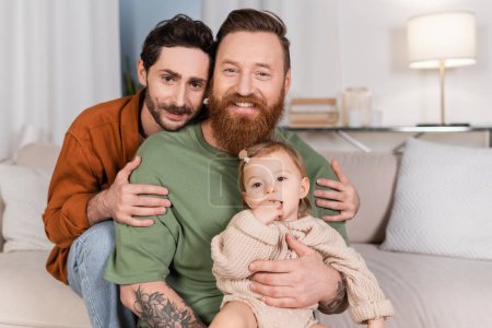 Foto de Sonriente gay hombre abrazando pareja con bebé hija en casa - Imagen libre de derechos