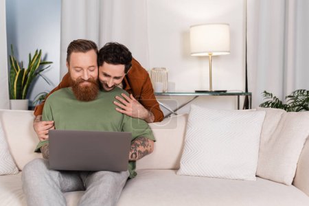 Foto de Gay hombre abrazando pareja usando laptop en sala de estar - Imagen libre de derechos