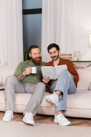 Photo for Positive gay couple with coffee reading book at home - Royalty Free Image
