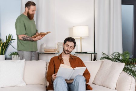 Gay man reading book near partner in living room at home 